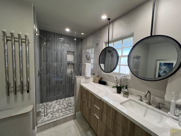 Shower with stacked grey wall tile and a pebble tiled floor, beside a wood vanity.