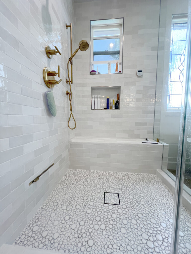 Inside a walk in shower, showing tiled walls, a recessed niche and a pebble floor with a square drain.