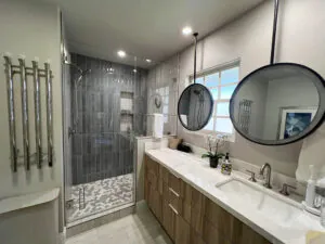 Double vanity with two round mirrors and a grey tiled shower with towel warmers