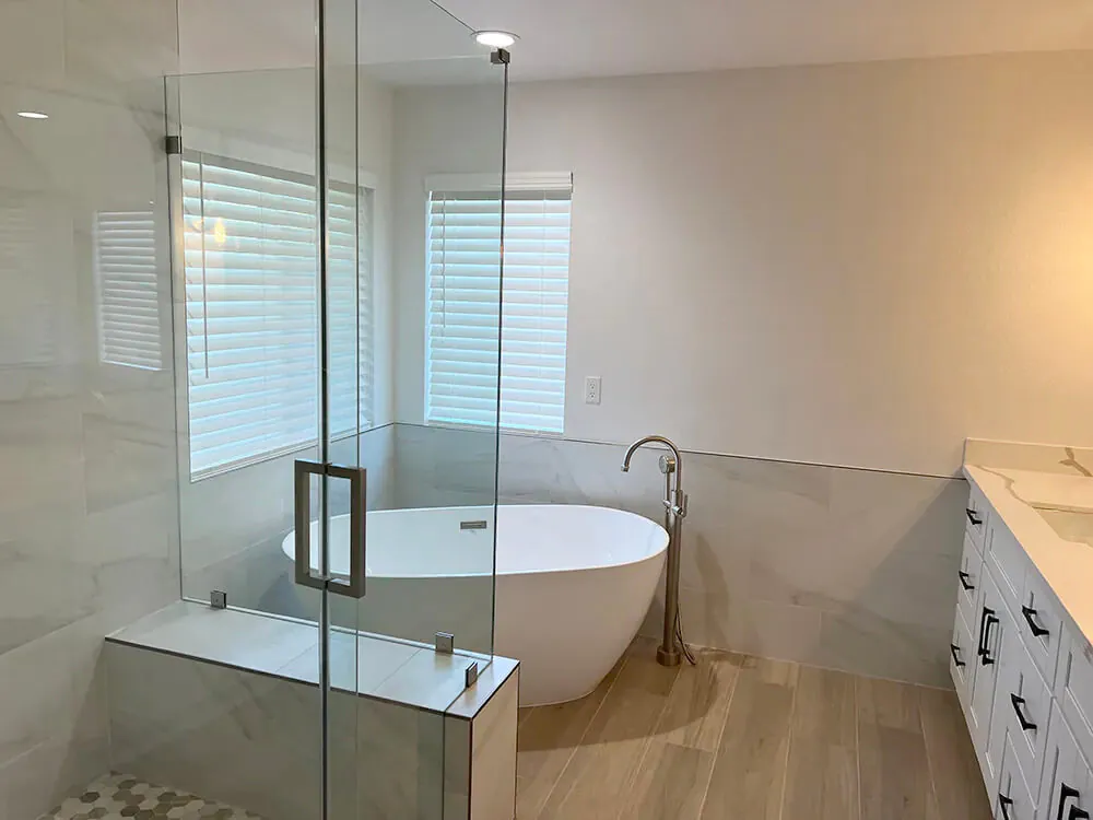Freestanding tub beside a glass walled shower, with a long vanity and plantation shutters